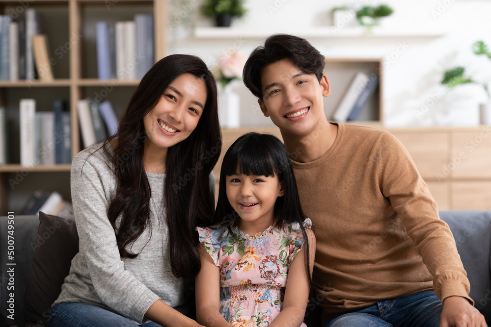 Happy Asian family sitting on couch smile with love and looking at camera spend time together.Father mother and little daughter relax on sofa in cozy living room.Good moment.Happy and warmth family. Happy Asian family sitting on couch smile with love and looking at camera spend time together.Father mother and little daughter relax on sofa in cozy living room.Good moment.Happy and warmth family.