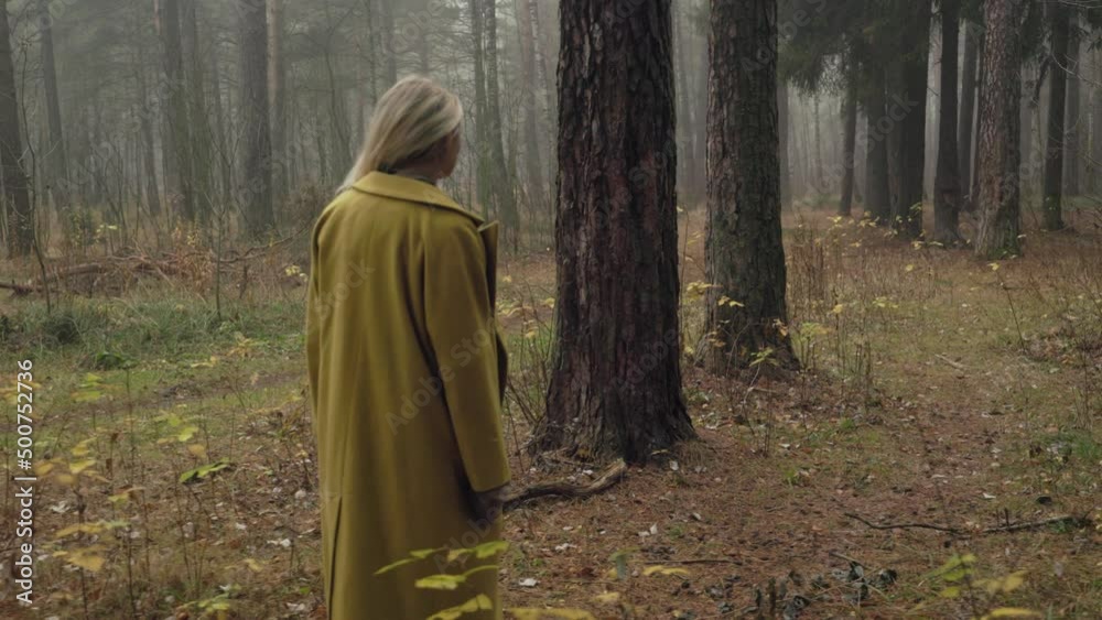 A young woman with dreadlocks in a yellow coat walks through a foggy autumn forest, side tracking shot