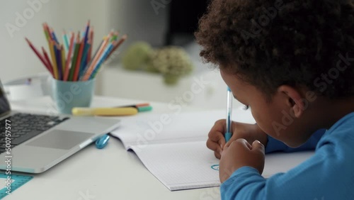 The African child with curly hair, learning with online primary school tutor lessons while at home sitting at the desk in modern apartmant, using a laptop, writing exercise book. Close-up. 4k footage