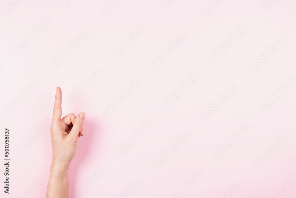 Woman hand pointing up with index finger to something virtual and invisible, copy space on pastel pink background. Hand gesture, number one