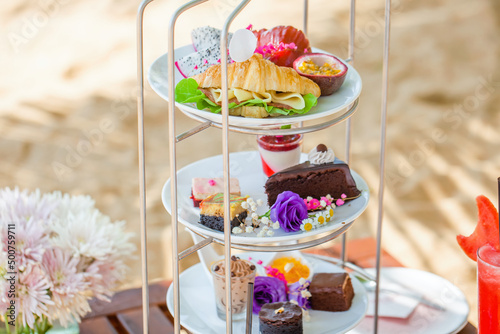 Afternoon tea on tropical sandy beach with shadows from palm trees in hot summer day. Cake stand with fresh pastries  sandwiches  sweeties  shake. Travel in Thailand  luxury hotel and restaurant