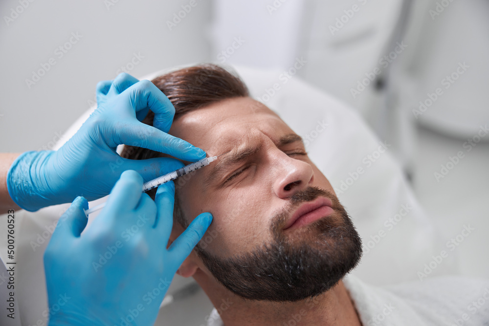 Ppatient grimaces of pain while careful cosmetologist does injection of lifting filler in his brow in clinic Ppatient grimaces of pain while careful cosmetologist does injection of lifting filler in his brow in clinic