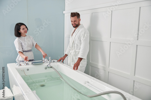 Specialist prepares hydro massage tub with clear water to mature guest in spa salon