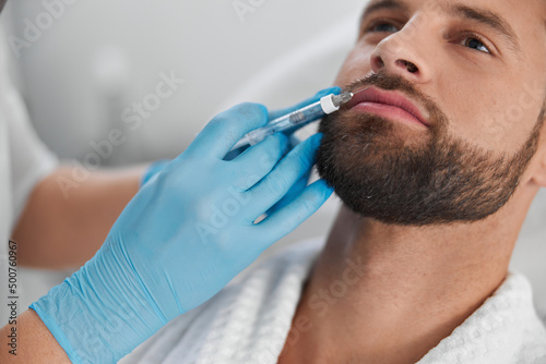 Fotografie, Obraz Attractive man with beard undergoes lip augmentation procedure with cosmetologis