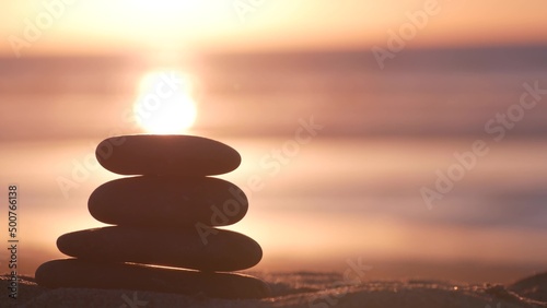 Stack of pebble stones  sandy ocean beach  sunset sky. Rock balancing in sun light  sea water waves. Stones staking in pyramid pile. Zen meditation and harmony in balance. Seamless looped cinemagraph.