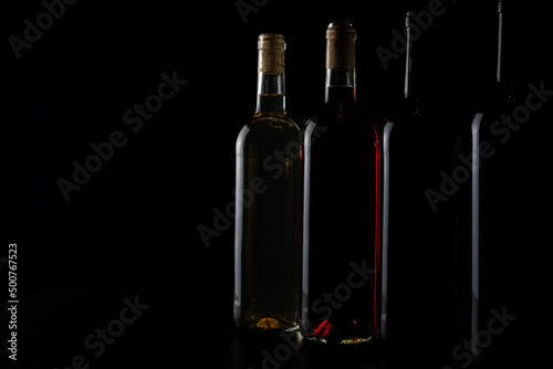 Several bottles of red, rosé and white wine in a row and diminishing perspective, with little light, revealing the relief of the glass, on a black background.