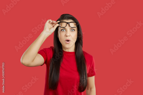 Beautiful young brunette woman impressed by something extraordinary looks at camera with surprised, shocked, astonished face expression, lifts her glasses and says WOW. Studio shot on red background photo