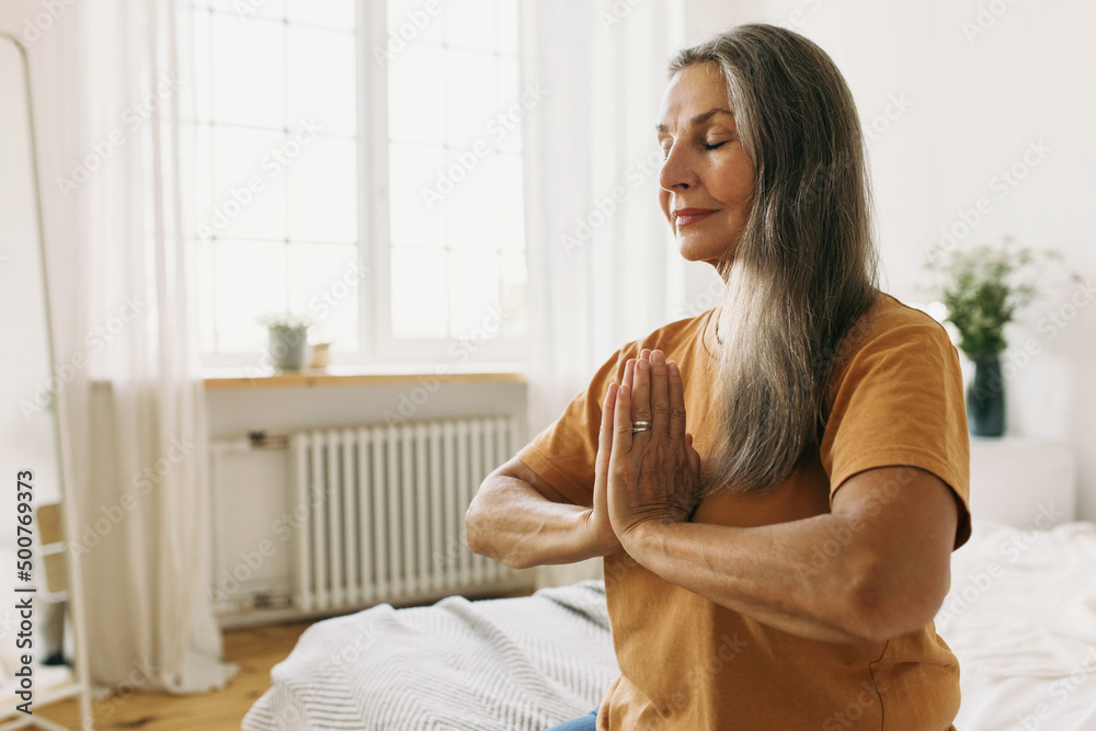 Elderly retired woman in good shape doing yoga exercises sitting at home on edge of her bed dressed in sporty yellow t-shirt having long grey dark blond hair, meditating with eyes closed Elderly retired woman in good shape doing yoga exercises sitting at home on edge of her bed dressed in sporty yellow t-shirt having long grey dark blond hair, meditating with eyes closed