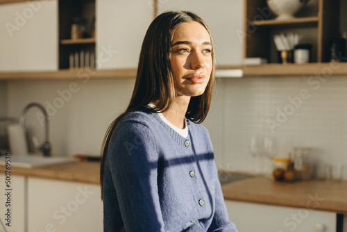 Profile picture of sun drenched beautiful female face against kitchen background, looking aside, wearing blue wool cardigan with buttons, having sensual pouty lips and long smooth hair