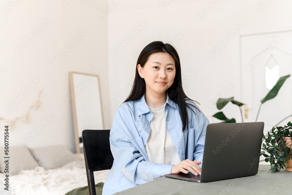 Young Asian woman working on a laptop at home and looking at the camera. Freelance business concept. Young Asian woman working on a laptop at home and looking at the camera. Freelance business concept.