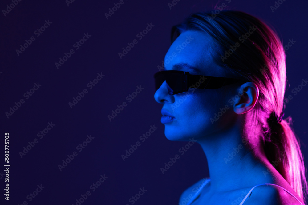 Young woman posing in a studio in a trendy neon light. Woman in sunglasses and hoodie with colorful light effects. Young woman posing in a studio in a trendy neon light. Woman in sunglasses and hoodie with colorful light effects.
