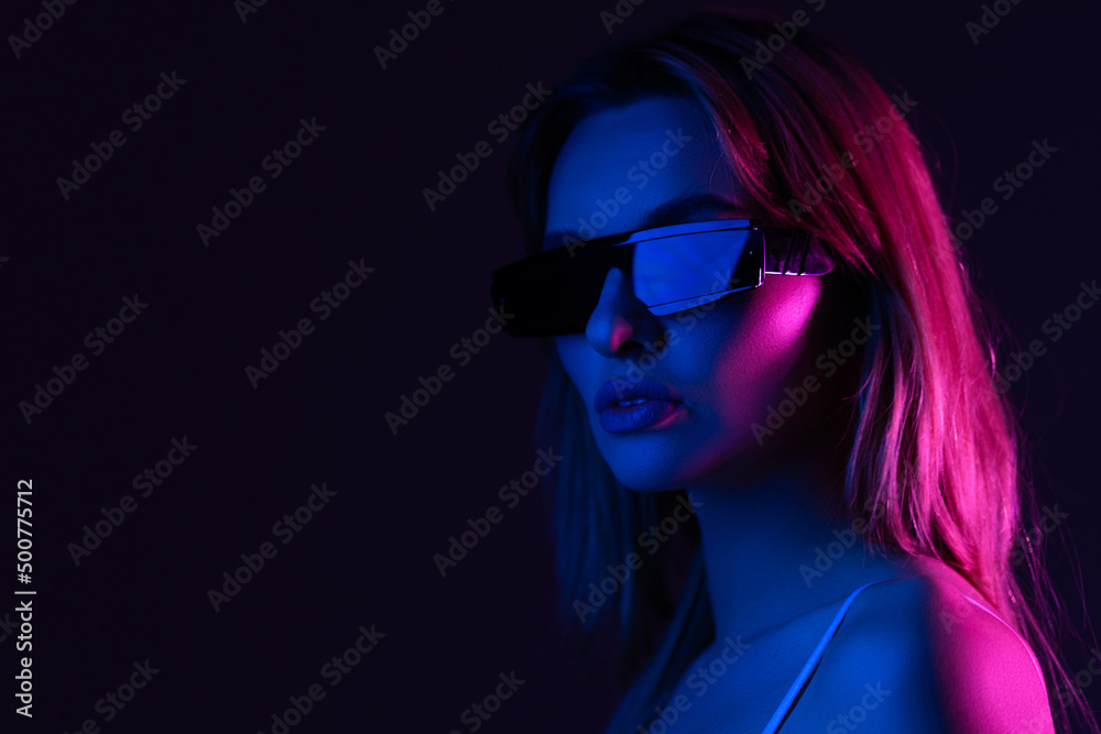 Young woman posing in a studio in a trendy neon light. Woman in sunglasses and hoodie with colorful light effects. Young woman posing in a studio in a trendy neon light. Woman in sunglasses and hoodie with colorful light effects.