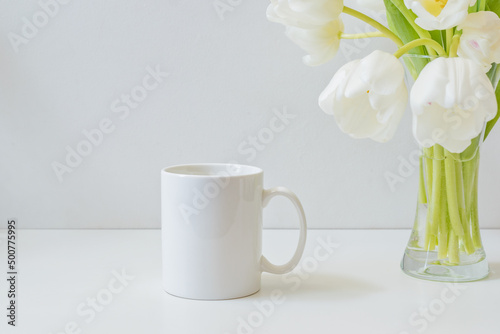 Mock up white mug and white spring tulips in a vase on a white table
