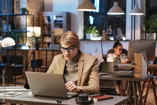Young Caucasian man in eyeglasses and jacket sitting at desk and using laptop while working In Dark Office
