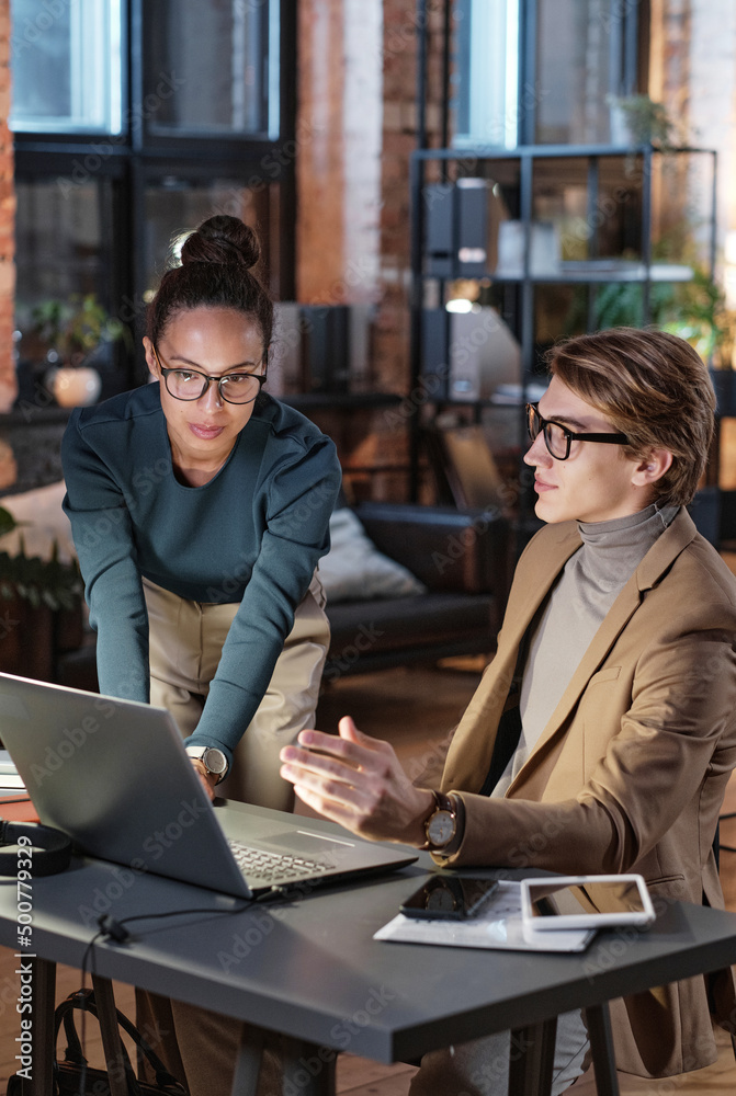 Female project manager with hairbun looking at laptop screen while analyzing internet resources with marketing specialist in dark office Female project manager with hairbun looking at laptop screen while analyzing internet resources with marketing specialist in dark office