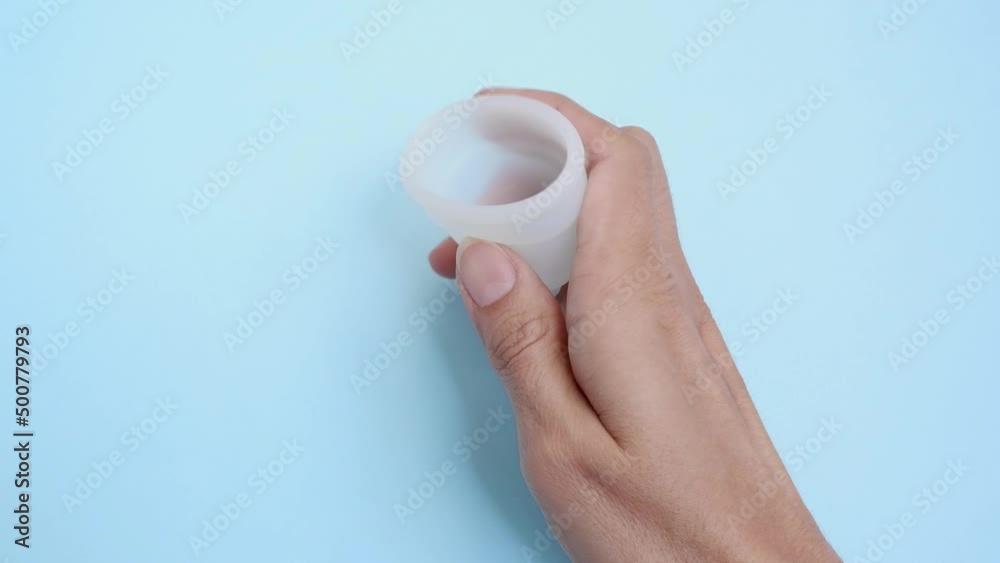 A woman holds a menstrual cup in her hands. White menstrual cup on a blue background. close-up of a hand showing a menstrual cup. Zero waste concept. An alternative feminine hygiene product in the per A woman holds a menstrual cup in her hands. White menstrual cup on a blue background. close-up of a hand showing a menstrual cup. Zero waste concept. An alternative feminine hygiene product in the per