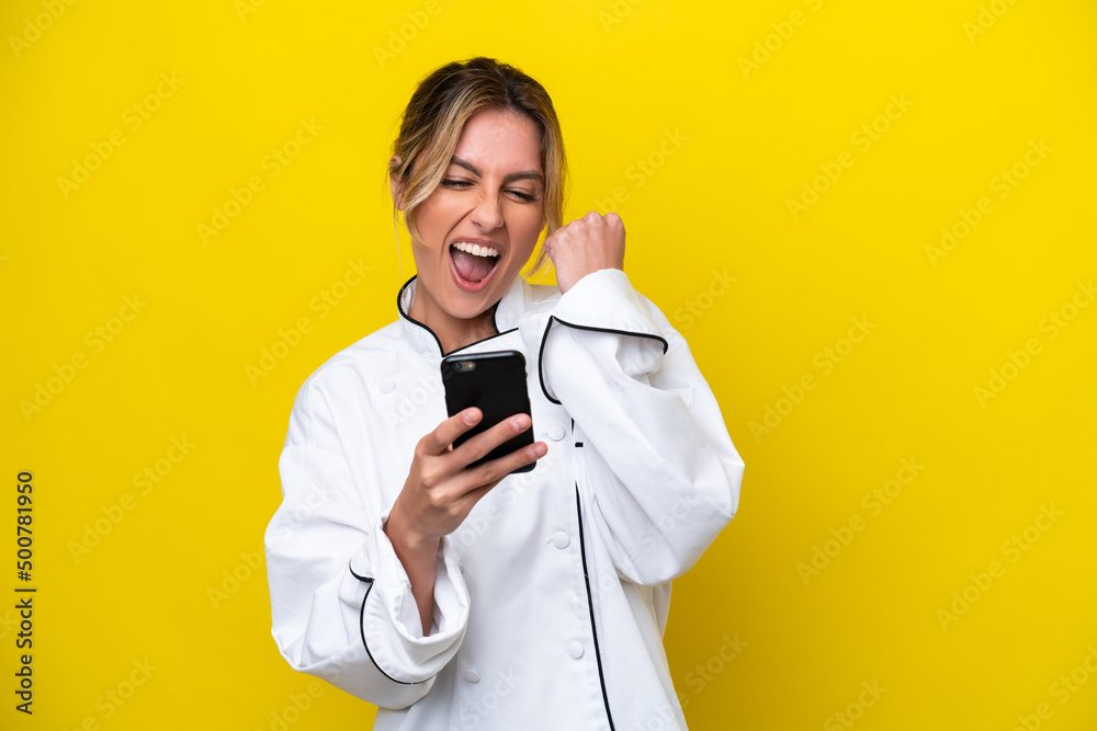 Uruguayan chef woman isolated on yellow background with phone in victory position Uruguayan chef woman isolated on yellow background with phone in victory position
