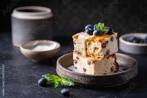 Baked homemade cottage cheese casserole or pudding with raisins serving with blueberry and sour cream on dark background. Close-up.