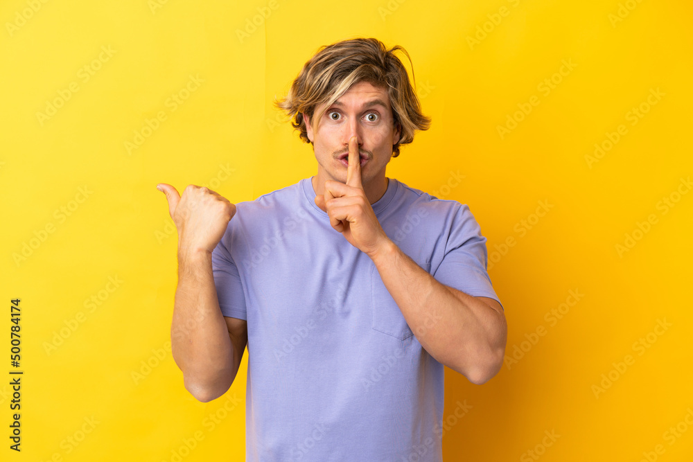 Handsome blonde man isolated on yellow background pointing to the side and doing silence gesture