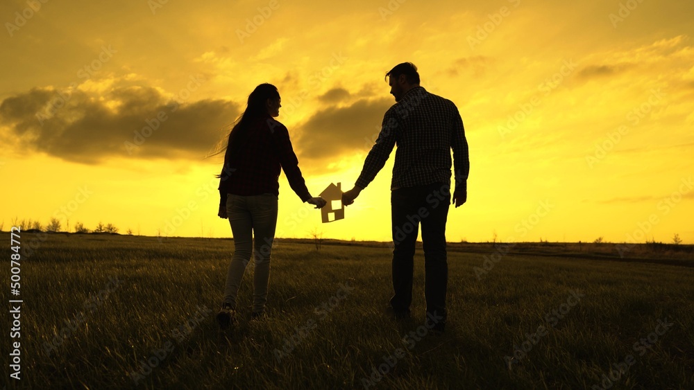 Family's hands are holding paper house at sunset, sun is shining through window. Symbol of house, happiness. Concept of building house for family. Dream to buy house. Family silhouette, family home