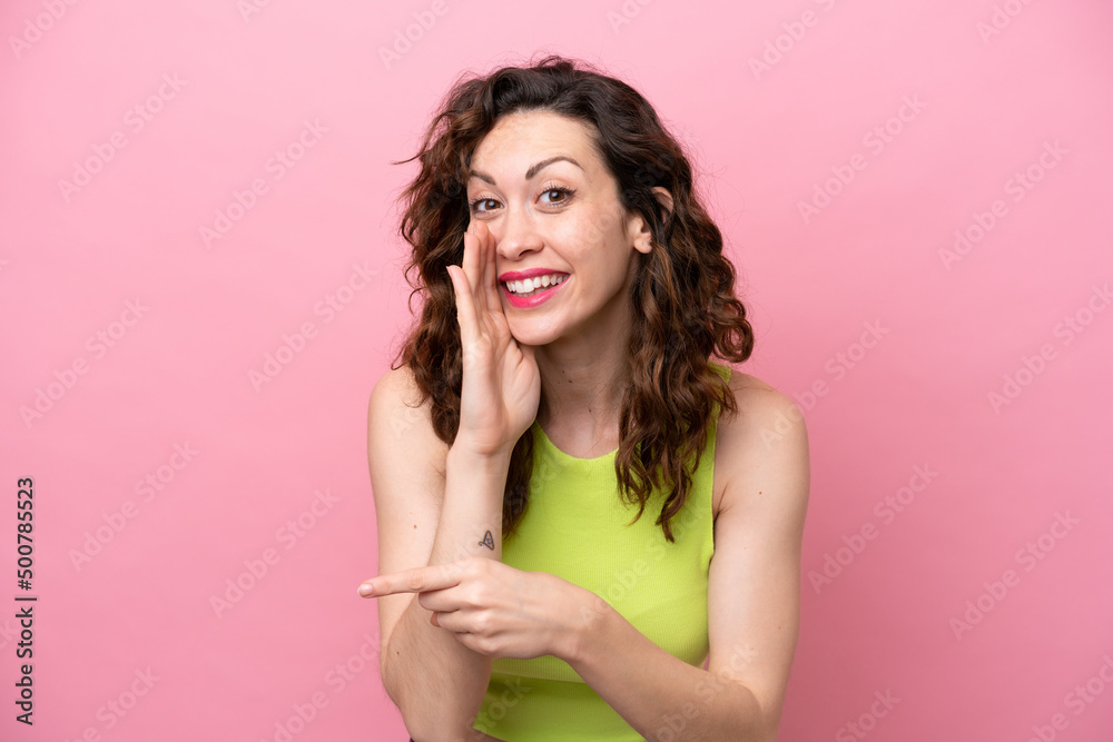 Young caucasian woman isolated on pink background pointing to the side to present a product and whispering something