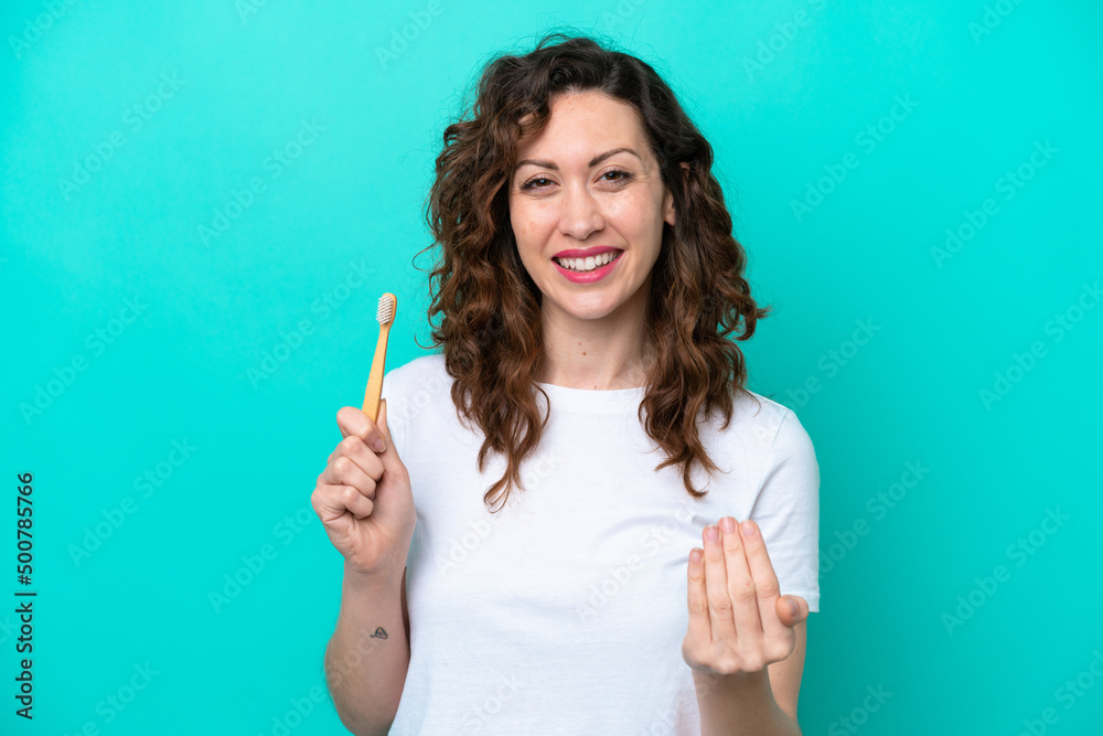 Young caucasian woman brushing teeth isolated on blue background inviting to come with hand. Happy that you came Young caucasian woman brushing teeth isolated on blue background inviting to come with hand. Happy that you came