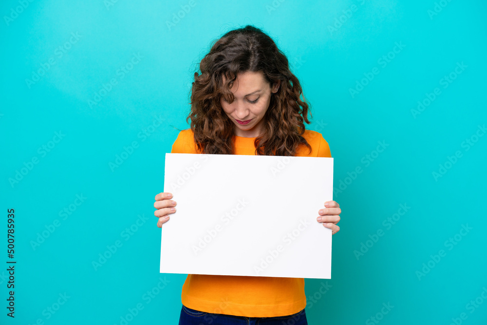 Young caucasian woman isolated on blue background holding an empty placard and looking it Young caucasian woman isolated on blue background holding an empty placard and looking it