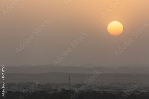 Sunset in Siwa. Yellow sky because of dust and sand in the air and silhouettes of local houses and a mosque. Layers of hills and mountains. Egypt