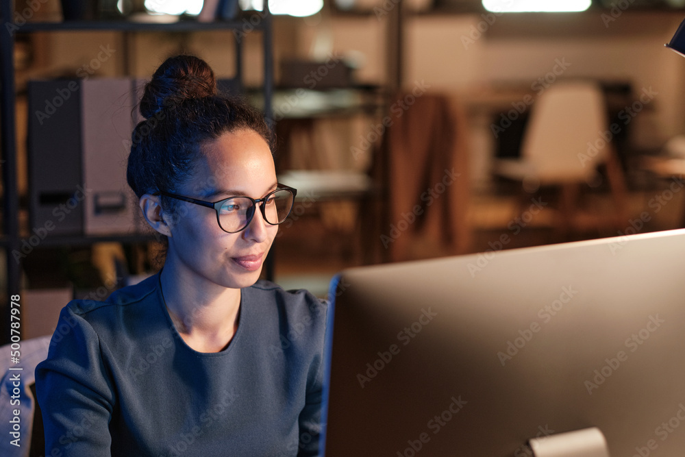 Content young biracial female office employee in glasses using computer while working at night