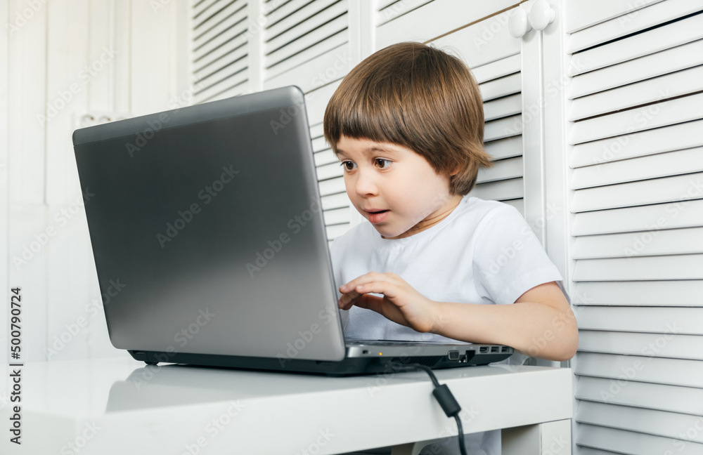 Boy sits at the table, uses laptop, typing on a keyboard, looks at the screen. Child does homework lesson, plays video game, study IT course. Home distance online education concept. Smart technology Boy sits at the table, uses laptop, typing on a keyboard, looks at the screen. Child does homework lesson, plays video game, study IT course. Home distance online education concept. Smart technology