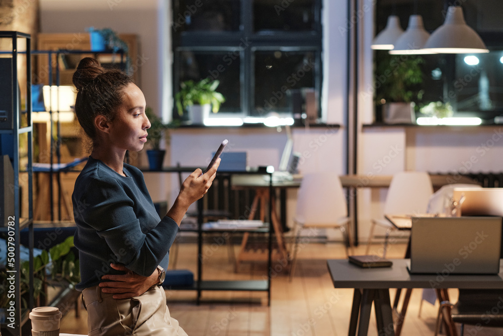 Serious attractive young mixed race businesswoman standing with smartphone in empty office at night