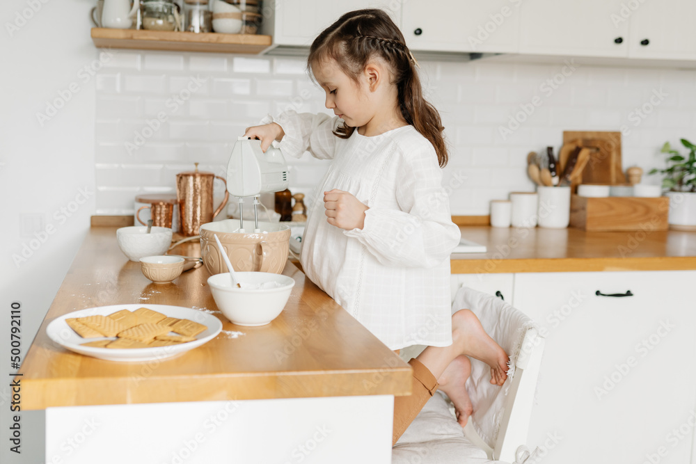 Little Caucasian brown-haired girl baking on white kitchen at home. Adorable girl preparing icing pancakes at the kitchen. Concept of food preparation.