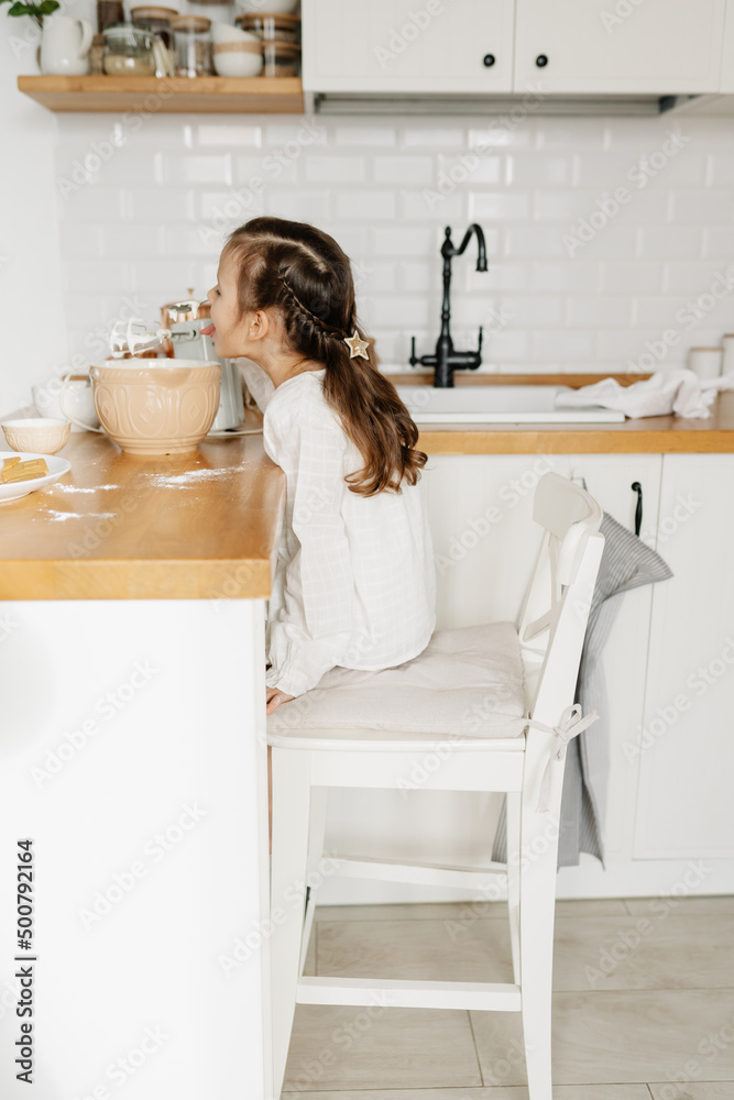 Little Caucasian brown-haired girl baking on white kitchen at home. Adorable girl preparing icing pancakes at the kitchen. Concept of food preparation. Casual lifestyle photo real life Little Caucasian brown-haired girl baking on white kitchen at home. Adorable girl preparing icing pancakes at the kitchen. Concept of food preparation. Casual lifestyle photo real life