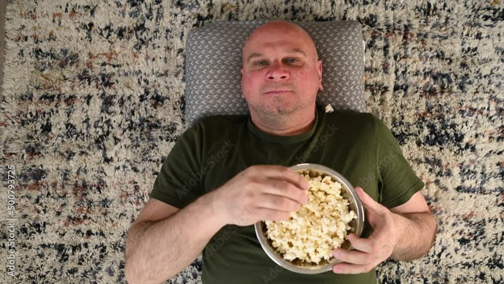 Man is getting bored waiting for a movie to start so he is starting to play a silly game with a bowl of popcorn. He is throwing up popcorn and trying to catch it with his mouth until losing his cool