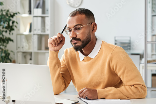 Serious busy young middle eastern marketer with beard holding pen and looking at laptop screen while working on strategy in office