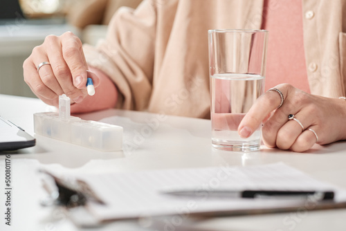 Close-up of unrecognizable businesswoman with glass of water taking pill from organizer at desk