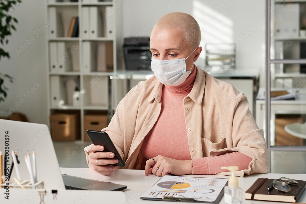 Concentrated young Caucasian bald woman in mask sitting with smartphone at table while working with business papers in office