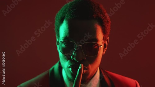 Stop talking. Gentleman secret. Confidential conspiracy. Neon light confident man in suit glasses warning with hush gesture isolated on dark red. photo