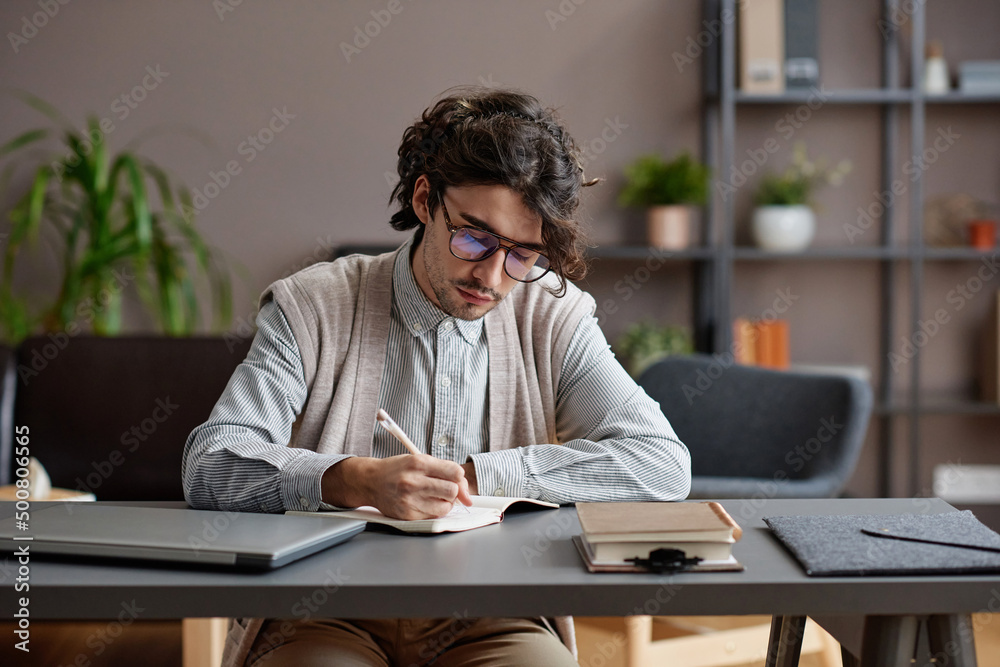 Horizontal medium portrait of young adult man wearing comfortable casual outfit and eyeglasses sitting at desk writing something in notebook Horizontal medium portrait of young adult man wearing comfortable casual outfit and eyeglasses sitting at desk writing something in notebook