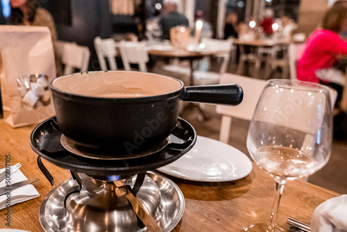 Heated pot of cheese fondue on burner. Wineglass and plates arranged on table. Gourmet swiss fondue dinner at luxurious ski resort during winter.