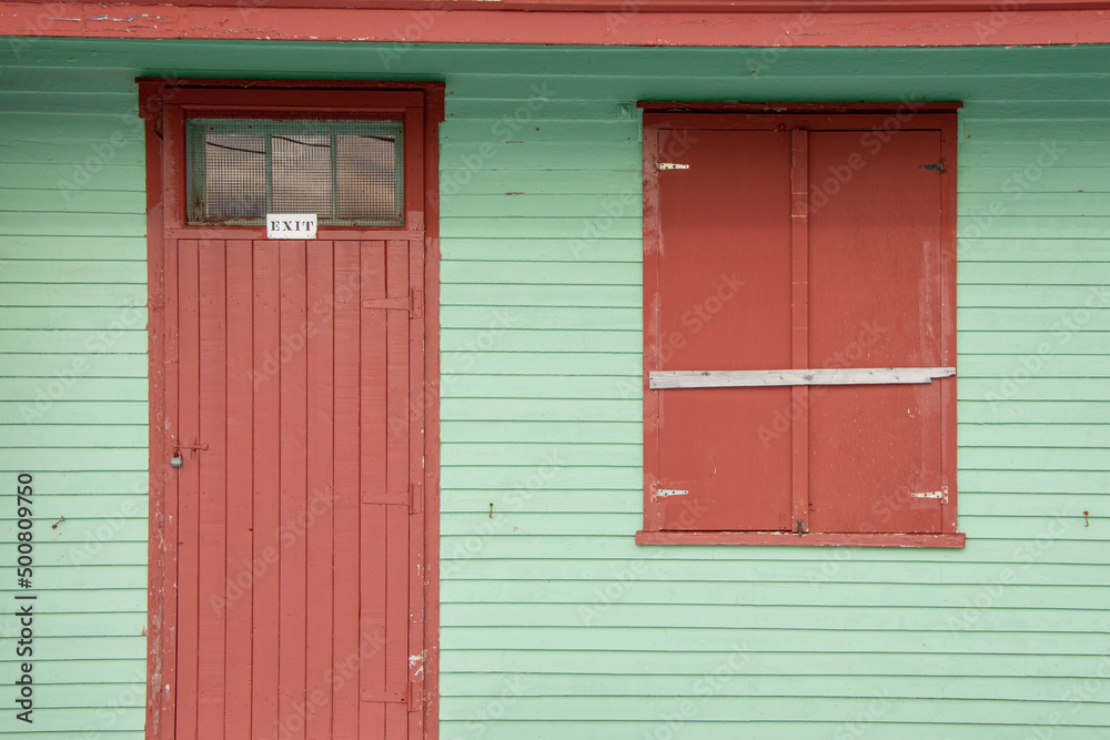 A vibrant mint green painted exterior of a building with a wooden shutter door and a closed double shutter covered window. There's a small white exit sign over the single door. The wall is lat siding A vibrant mint green painted exterior of a building with a wooden shutter door and a closed double shutter covered window. There's a small white exit sign over the single door. The wall is lat siding