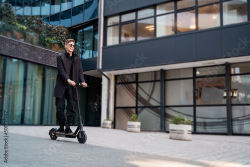 Young , elegant man in coat driving e scooter through the city, modern building in the back 