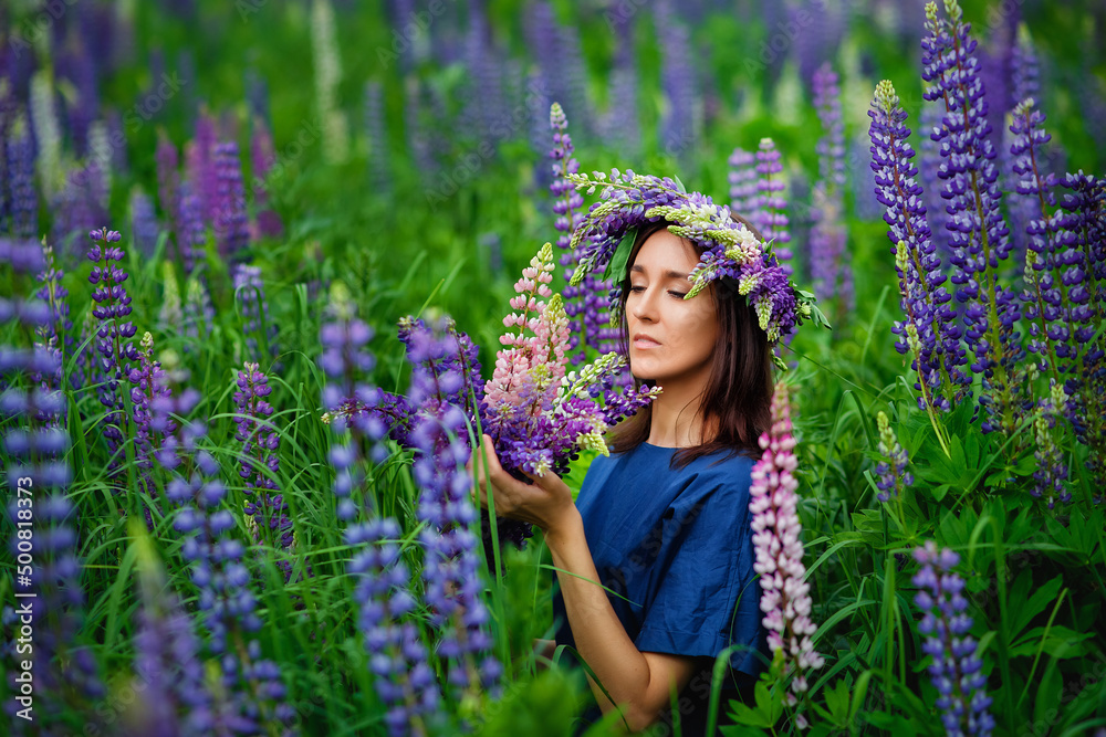 Fototapeta premium A young brunette woman in a flower wreath made of lupine with a bouquet of lupines smiles gently and looks at the camera in a flower meadow, inhales the fragrance of flowers.