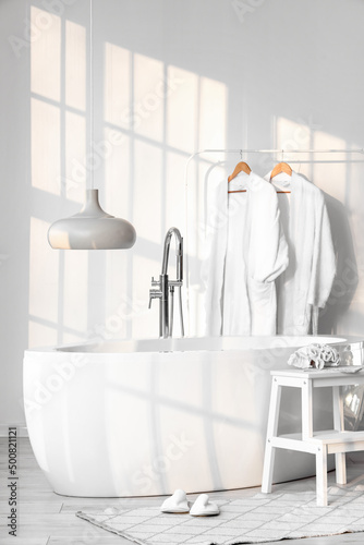 Stylish bathtub, rack with bathrobes, mirror and stepladder stool in room interior © Pixel-Shot