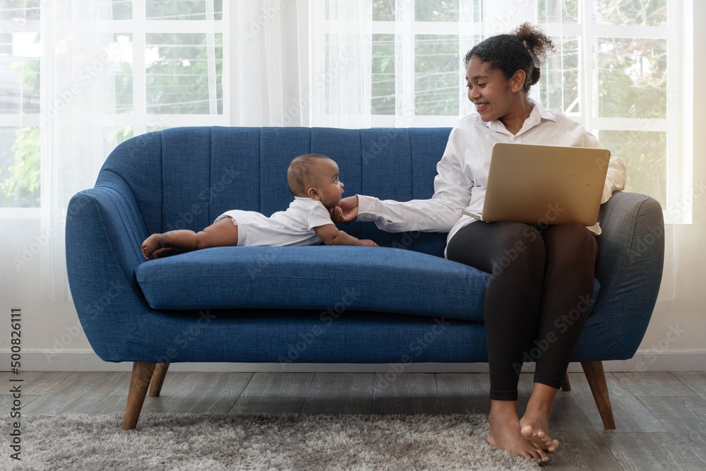 African mother working remotely on laptop while taking care of her baby on couch, freelance single mom working at home African mother working remotely on laptop while taking care of her baby on couch, freelance single mom working at home
