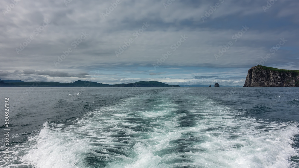 The foam trail from the boat on the surface of the water. The hills and rocks of Kamchatka are visible on the horizon. Cloudy sky. Pacific ocean. Avacha Bay.