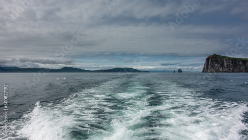 The foam trail from the boat on the surface of the water. The hills and rocks of Kamchatka are visible on the horizon. Cloudy sky. Pacific ocean. Avacha Bay.