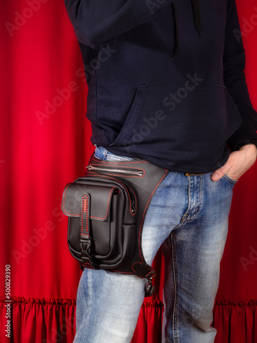 Bag on the belt, hip handmade of genuine black leather stitched with red thread. Bag on the hip of a man wearing jeans on a red background.