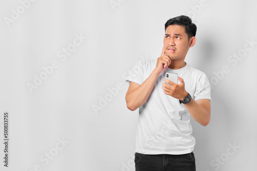 Pensive young Asian man with a serious face thinking about a question, using a mobile phone isolated on white background