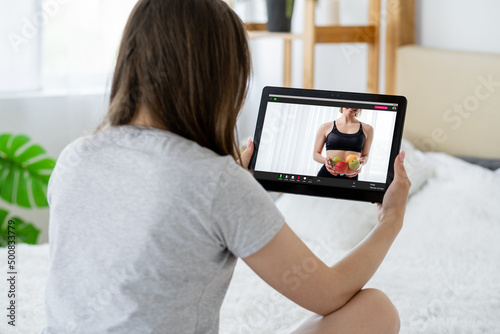 Online fitness diet. Weight loss app. Personal trainer video call. Unrecognizable woman watching athletic coach with fruit bowl on tablet screen at home interior.